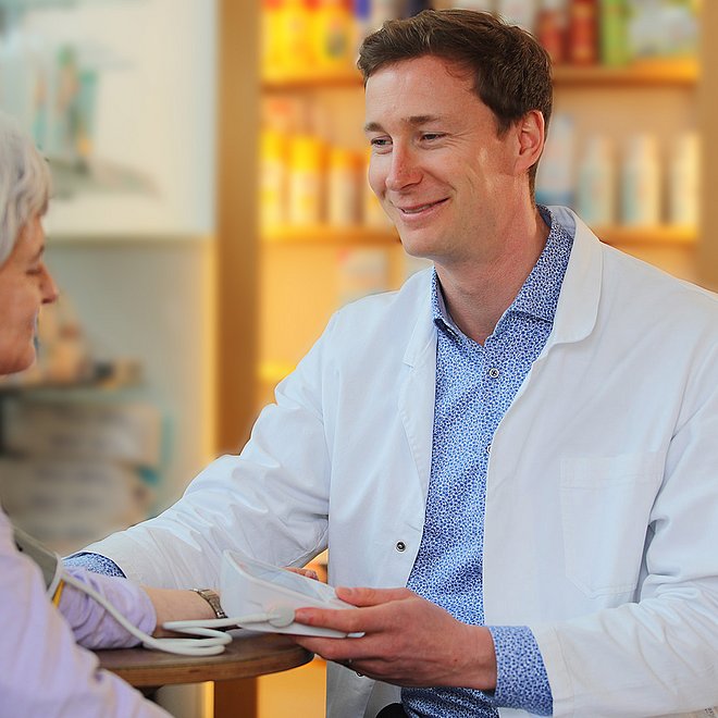 Ein Apotheker der BLAK misst bei einer älteren Dame den Blutdruck. Dabei sitzen Sie an einem Tisch. Hinter ihnen stehen die Medikamentenregale.