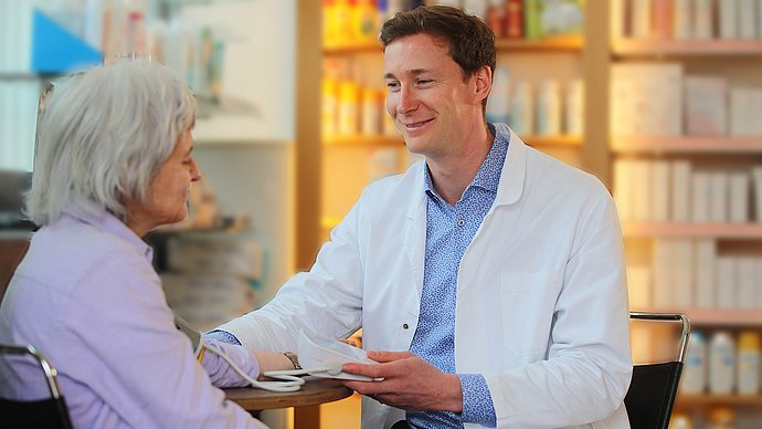 Ein Apotheker der BLAK misst bei einer älteren Dame den Blutdruck. Dabei sitzen Sie an einem Tisch. Hinter ihnen stehen die Medikamentenregale.